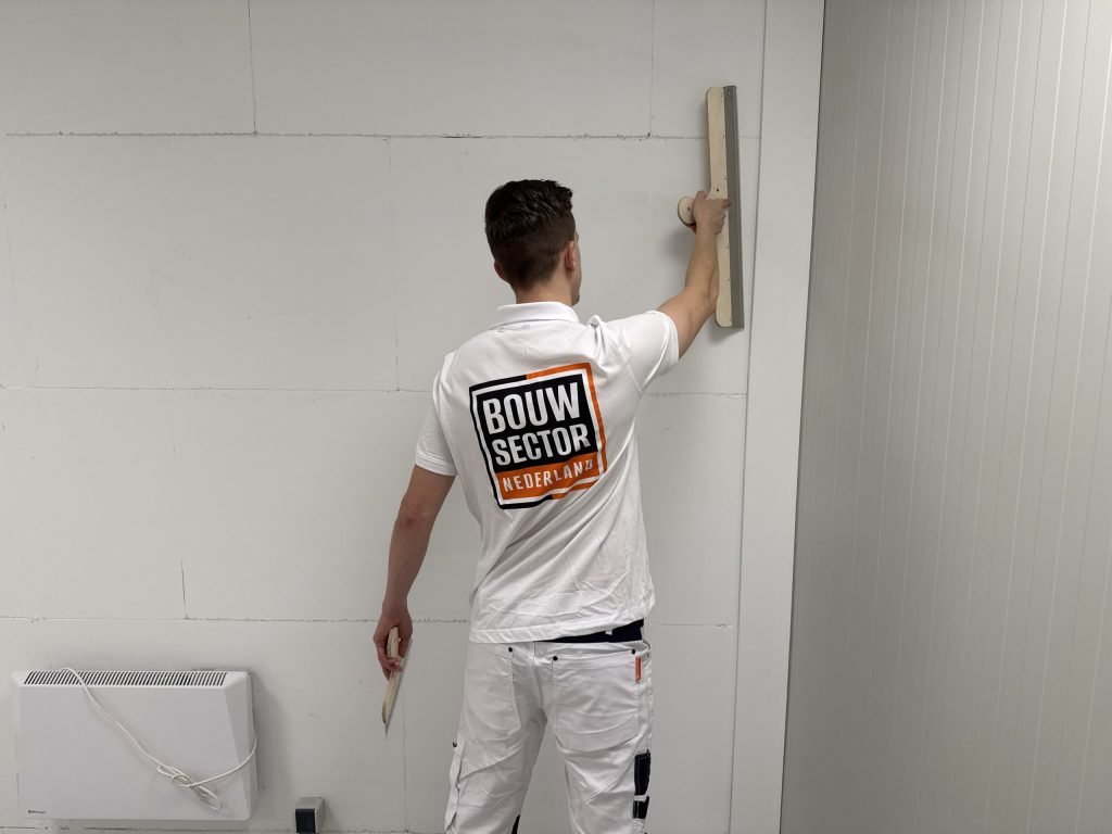 Stukadoor met het Bouwsector-logo op zijn rug zet een lange spaan rechtop tegen de bovenkant van een wand