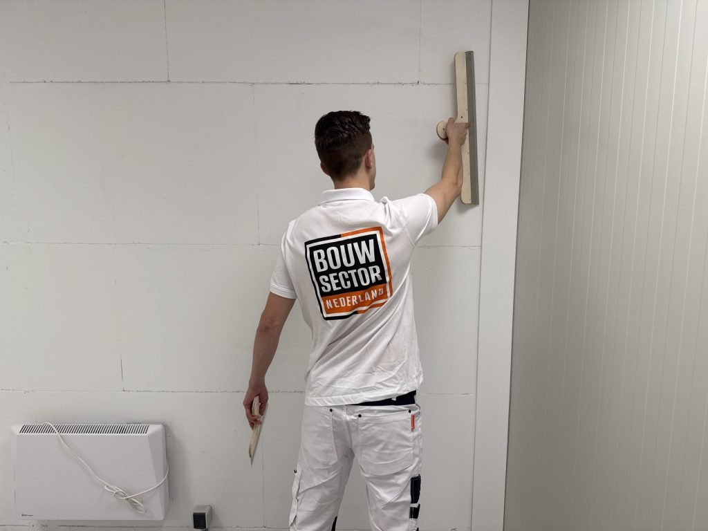 Stukadoor in witte werkkleding houdt een brede spaan hoog tegen een wand, naast een elektrische kachel
