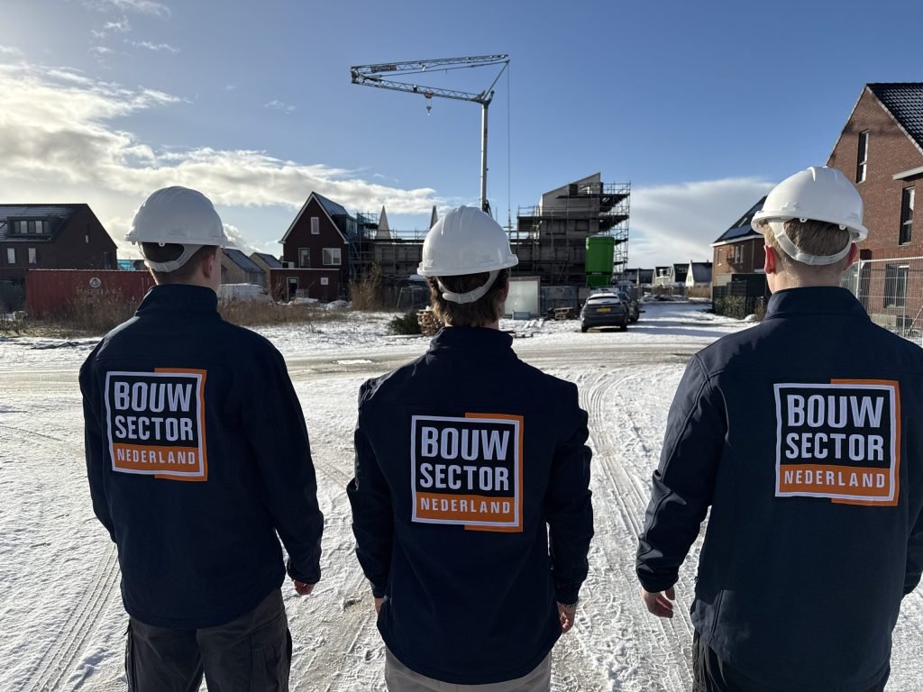 Drie bouwvakkers van Bouwsector Nederland kijken naar een rij woningen in aanbouw onder een blauwe winterlucht