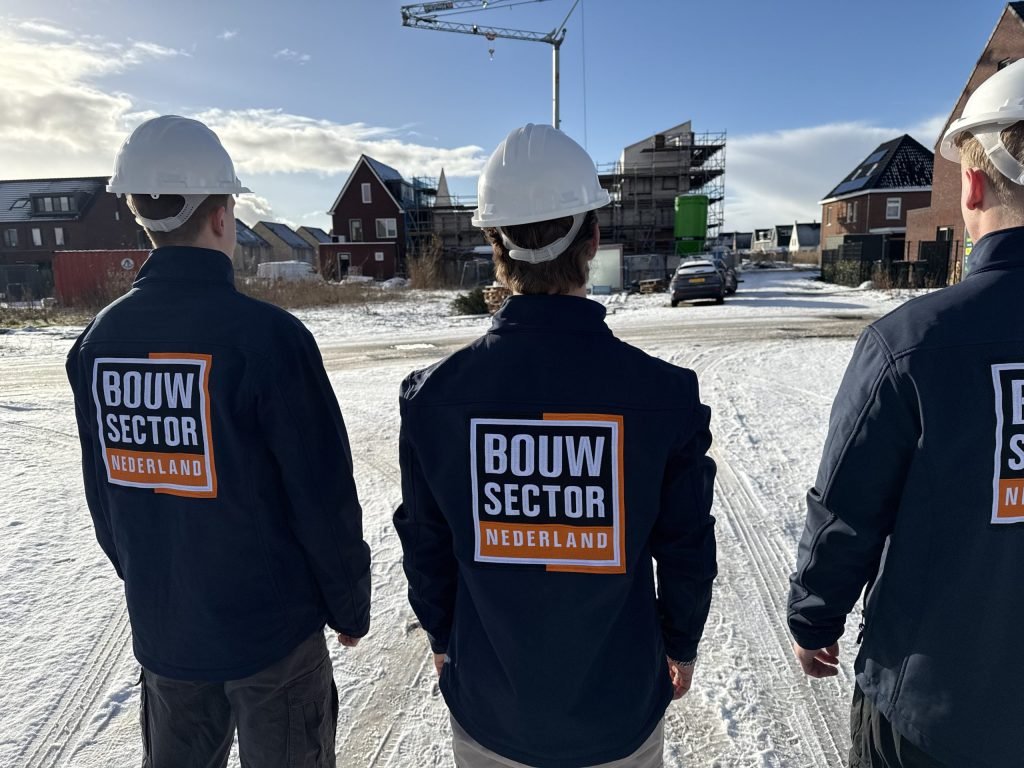 Drie vakmensen van Bouwsector Nederland in blauwe jassen naast elkaar op een bouwplaats met een hijskraan